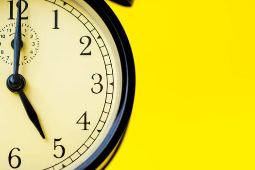 Closeup of a vintage analog black clock on a yellow background displaying 5 o'clock