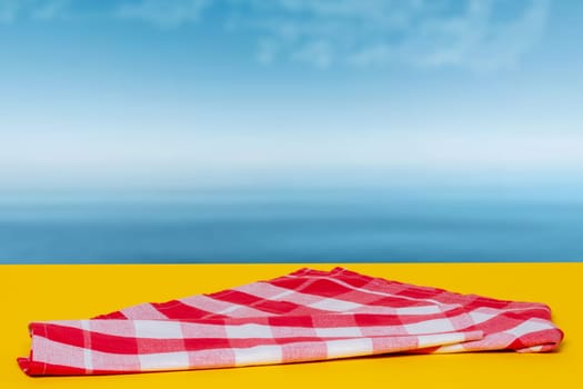 Empty table product. Empty red checkered tablecloth on yellow table over abstract blurred ocean background. Space for your food and product display montage.