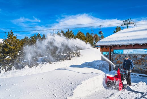 El Tarter, Andorra, Jan 2020 Men operating hand pushed wheel snow plow and cutting trough snowdrift, shooting up snow powder, Pyrenees Mountains