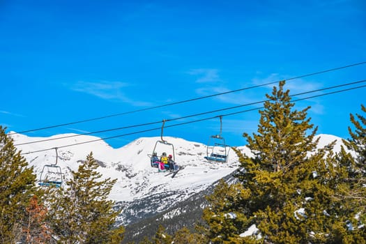Group of people, skiers riding up on chair ski lift with snowy mountains and forest in a background. Winter holidays in Andorra, Pyrenees Mountains