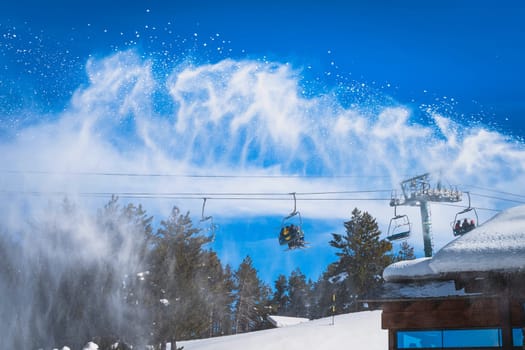 Snow cannon spreading fresh snow powder on a ski slope. Skiers riding up on chair lift. Winter ski holidays in El Tarter, Andorra, Pyrenees Mountains