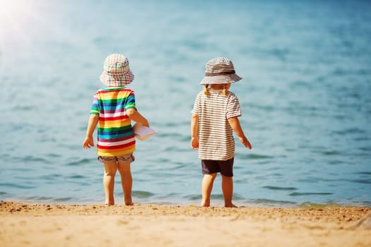 Girl and boy with paper boat in his hands going to the sea water along shore. Concept of the family vacation, childhood and friendship.