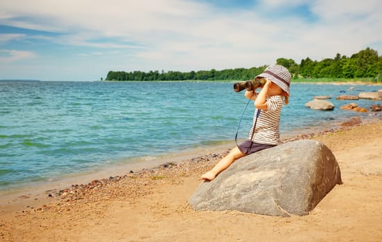 Little girl sitting on the stone on the seashore with binocular in her hands. Concept of the childhood and family vacation.
