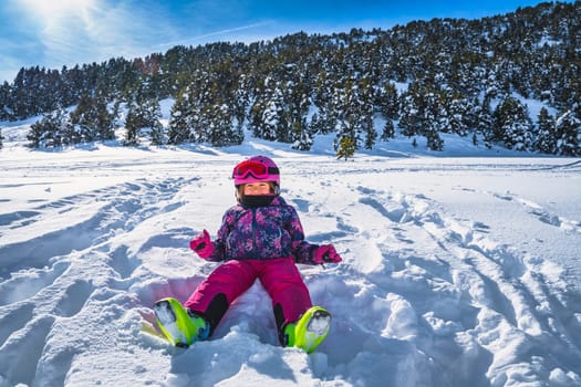 Smiling girl sitting on fresh snow powder in a ski gear with mountains and forest in a background. Winter ski holidays, Andorra, El Tarter, Pyrenees