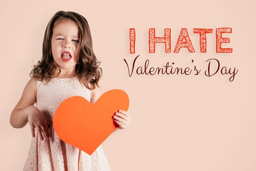 Portrait of little girl with long curly hair hold in hand big red paper heart showing disgust on peach background. Inscription I hate Valentines Day. Post or Greeting card. Copy space
