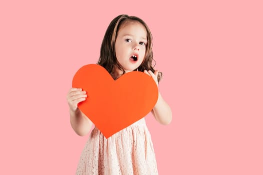Little girl with long curly hair hold in hand big red paper heart opening mouth surprisingly isolated on pink background. Congratulations, Valentines day, birthday, Mother's day concept. Copy space.