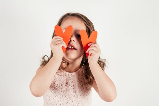 Portrait of beautiful little girl with long curly hair hold in hands two red paper hearts covering eyes on white background. Family, love, holidays, birthday, Mother's day concept. Copy space. Mock up