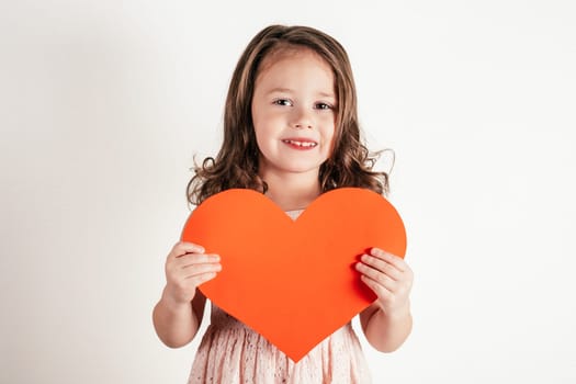 Portrait of little girl with long curly hair hold in hands red paper heart smiling on white background. Congratulations on family holidays, Valentines day, birthday, Mother's day concept. Copy space.