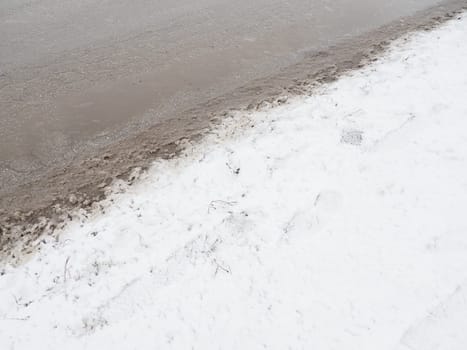Roadside with a snowdrift. Difficult driving conditions. Wet slippery pavement with footprints. Dangerous ice. Muddy weather in winter. Car tire tracks. The work of municipal services for cleaning.