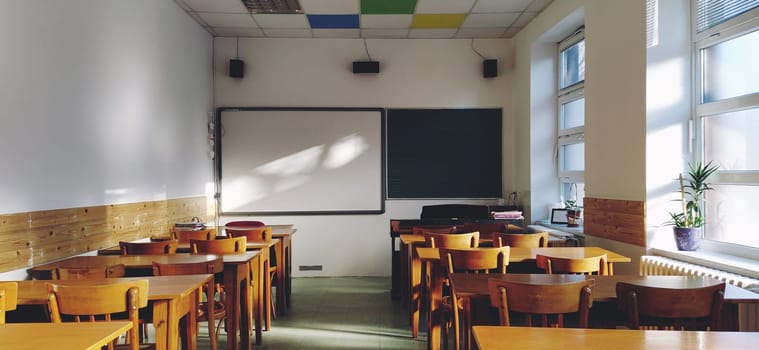 Music solfeggio empty class at school. The sun's rays fall on the floor through the window. Wooden student tables and chairs. School board and white walls. School interior