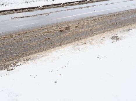 Roadside with a snowdrift. Difficult driving conditions. Wet slippery pavement with footprints. Dangerous ice. Muddy weather in winter. Car tire tracks. The work of municipal services for cleaning.