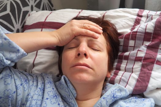 Sick woman in bed. Headache, runny nose and cold. Paleness of the face and faintness of the body. Feeling unwell. The patient holds his forehead with his hand and covers his eyes.