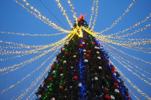 Multi-colored glass shiny balls on the New Year's city tree. Glowing garlands hang over the Christmas tree. Outdoor winter decoration. Festive design of the city square. Red, blue and yellow ball.