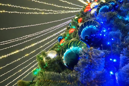 Multi-colored glass shiny balls on the New Year's city tree. Glowing garlands hang over the Christmas tree. Outdoor winter decoration. Festive design of the city square. Red, blue and yellow ball.