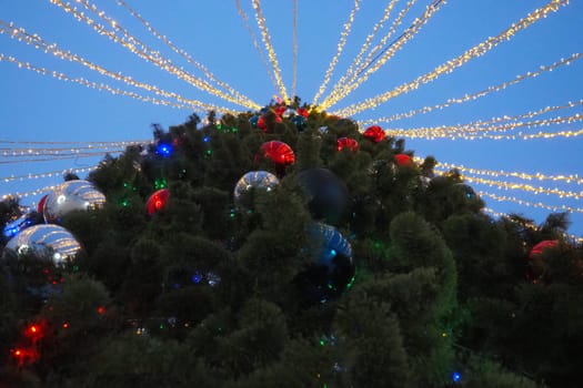 Multi-colored glass shiny balls on the New Year's city tree. Glowing garlands hang over the Christmas tree. Outdoor winter decoration. Festive design of the city square. Red, blue and yellow ball.