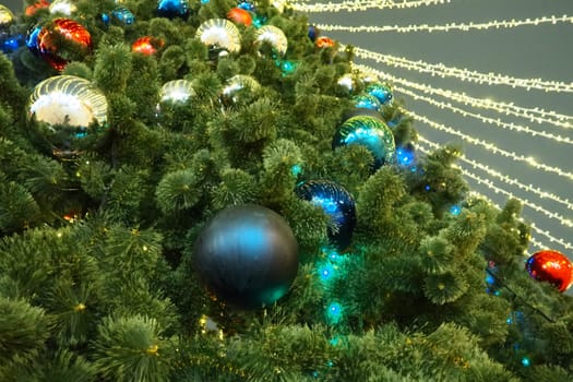 Multi-colored glass shiny balls on the New Year's city tree. Glowing garlands hang over the Christmas tree. Outdoor winter decoration. Festive design of the city square. Red, blue and yellow ball.
