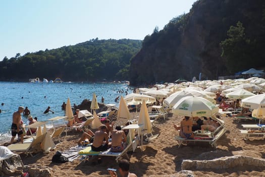 Budva, Montenegro, Mogren beach. August 15 2022 Sand and pebble beach near high cliffs. Beach umbrellas, cafes, deck chairs. People relax, swim, enjoy and sunbathe. Tourism business in the Balkans