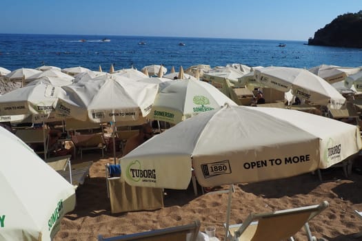 Budva, Montenegro, Mogren beach. August 15 2022 Sand and pebble beach near high cliffs. Beach umbrellas, cafes, deck chairs. People relax, swim, enjoy and sunbathe. Tourism business in the Balkans