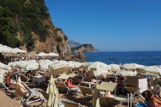 Budva, Montenegro, Mogren beach. August 15 2022 Sand and pebble beach near high cliffs. Beach umbrellas, cafes, deck chairs. People relax, swim, enjoy and sunbathe. Tourism business in the Balkans
