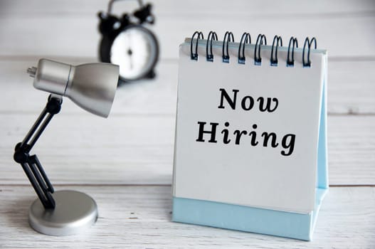 Now hiring text on notepad with table lamp on wooden background. Employment concept