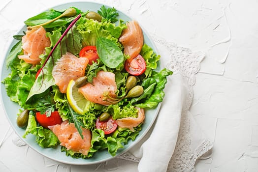 Fresh green salad with smoked salmon, cherry tomatoes and capers on white table background close up