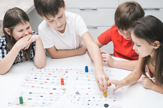 Top view of group of children friends classmates sitting at white table, playing board game tabletop game, moving colorful figures on white board with numbers, tracks. Board games, entertainment.
