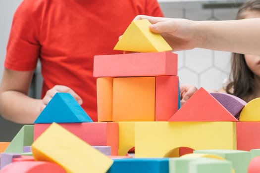 Cropped photo of preteen children friends sitting at table in kitchen, building tower house castle of colorful wooden construction blocks geometric figures. Board games, entertainment, development.