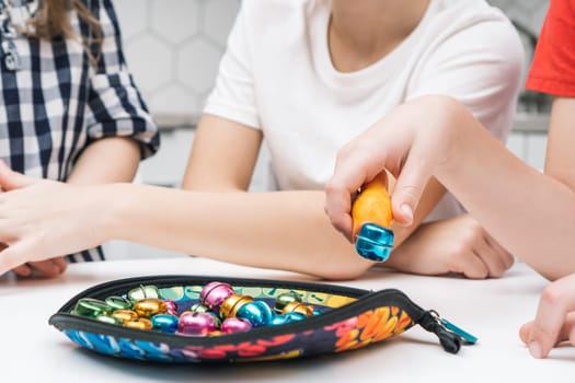 Side view of group of preteen children sitting at white table, catching colorful round metal bells with orange plastic tweezers on soft bright textile tray. Board game, entertainment, development.