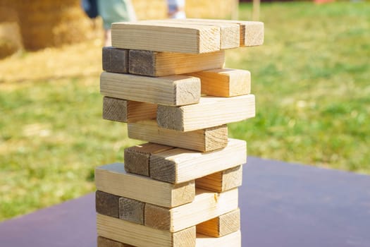 Side view of board game Jenga tower made of wooden blocks beams details built on blue table outdoors in park field in summer in sunny day. Board games, development, hobby, entertainment, education.