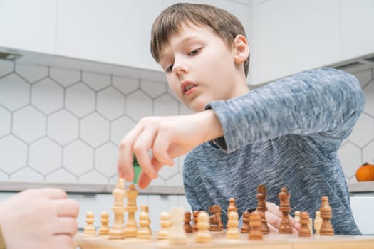 Portrait of serious preteen schoolboy sitting at table in white kitchen at home, playing chess on wooden board, planning, building strategy, taking light figure with hand. Board games, development.