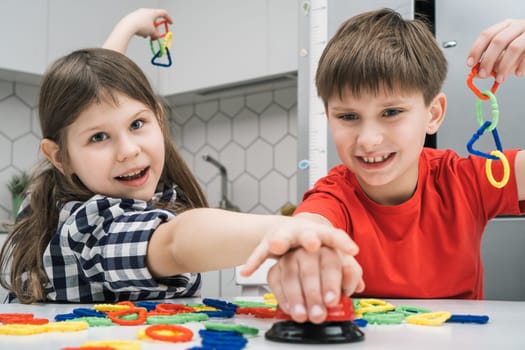 Portrait of two happy friends children sitting at table in kitchen at home, playing board game ring, pushing bell, joining different colorful geometric shapes figures in chain, having fun. Board game.