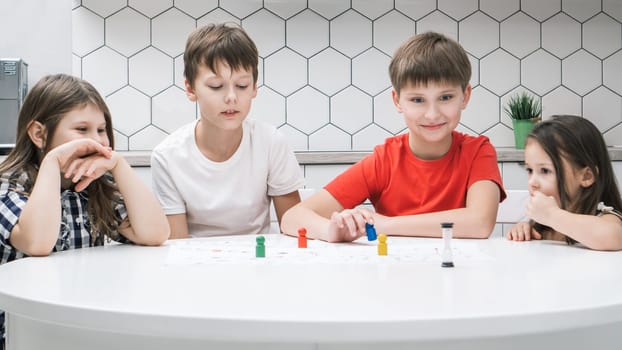 Portrait of group of excited children friends sitting at white table in kitchen at home, playing tabletop game, moving colorful figures on white board with numbers, tracks. Board games, entertainment.