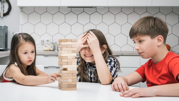 Group of focused children playing board game Jenga on table in kitchen at home building tower made of wooden blocks beams. Worried girl cover eyes with hands waiting friend pull detail. Entertainment.