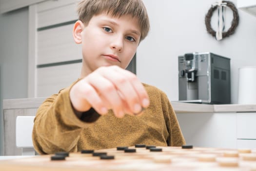 Portrait of serious preteen schoolboy sitting at table in kitchen at home, playing checkers on wooden board, planning, building strategy, holding black checker with hand. Board games, entertainment.