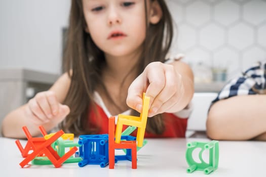 Cropped photo of preteen children sitting at white table in kitchen, playing board game stacking chairs, putting colorful small chairs in stack for building balancing tower. Board games. Soft focus.