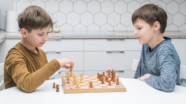 Side view of two calm focused preteen boys friends brothers sitting at white table in kitchen at home, playing chess on wooden board, competing. Board games, development, education, leisure, hobby.
