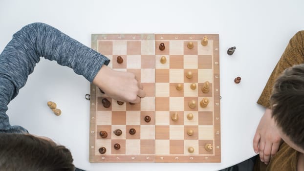 Top view of two focused preteen boys friends brothers sitting at white table, playing chess on wooden board, thinking, building strategy, holding figure with hand. Board games, development, education.