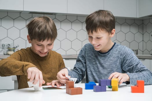 Portrait of children friends sitting at table in kitchen at home, reading instruction for playing colorful toy wooden construction blocks. Board games, entertainment, development, education, tetris.