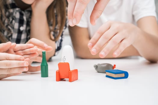 Cropped photo of group of children playing board game card game geistes blitz on white table in kitchen at home, choosing tiny figure: green bottle, red armchair, blue book, grey mouse, white ghost.