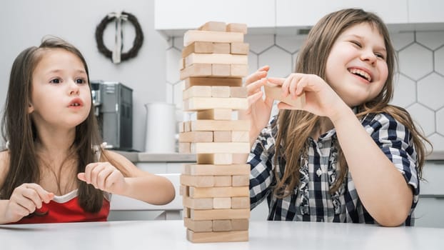 Two preteen girls friends children playing board game Jenga on table in kitchen at home, building tower made of wooden blocks, looking aside, laughing. Board games, hobby, entertainment, education.