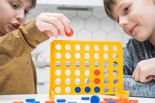 Portrait of two preteen boys friends children sitting at white table in kitchen at home, playing board game connect four 4 in line, inserting checkers into yellow plastic grid. Board games, education.