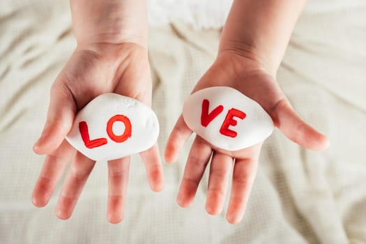 Hands of little girl hold two white smooth stones with syllables lo and ve painted red on white cloth background. Family, love, holidays, birthday, Valentine's day, Mother's day concept. Copy space.