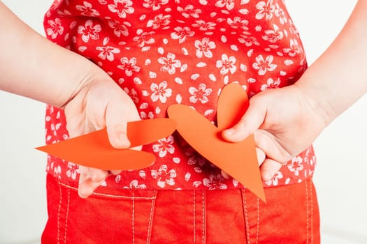 Portrait of little girl in red clothes hold in hands two red paper hearts together behind back on white background. Family, love, holidays, birthday, Valentine's day, Mother's day concept. Copy space.
