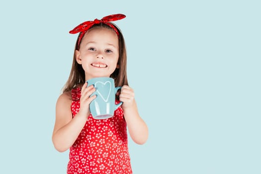 Portrait of little girl with long hair hold in hands blue mug with heart smiling isolated on blue background. Congratulations on Valentines day, birthday, Mother's day concept. Copy space.