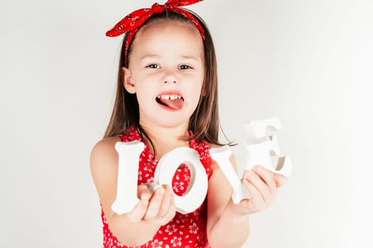 Little girl with long hair in red clothes hold white volumetric letters of word love in hands showing tongue on white background. Family, love, holidays, birthday, Mother's day concept. Copy space.