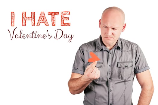 Handsome frowning bald middle-aged man in gray shirt hold red paper heart with two fingers and inscription I hate Valentines day isolated on white background. Valentine's day, love, emotion concept