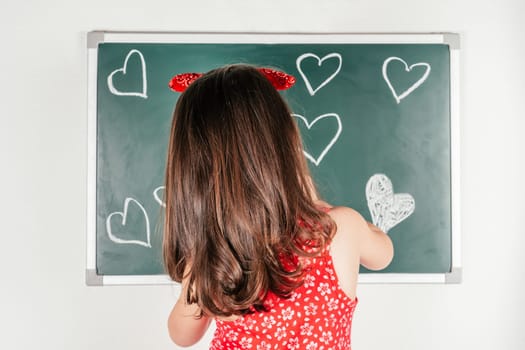 Little girl with long curly hair in red clothes standing back, drawing hearts with chalk on green blackboard on white background. Family, love, holidays, birthday, Mother's day concept. Copy space.