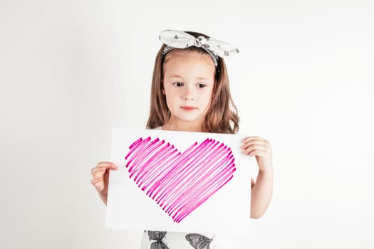 Portrait of little thoughtful girl with long hair holding in hands sheet of paper with pink heart on white background. Congratulations on family holidays, birthday, Mother's day concept. Copy space.
