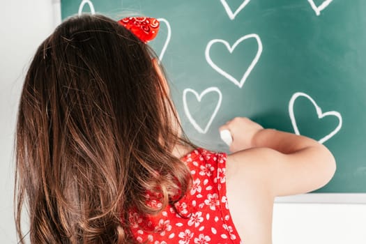 Little girl with long curly hair in red clothes standing back, drawing hearts with chalk on green blackboard on white background. Family, love, holidays, birthday, Mother's day concept. Copy space.