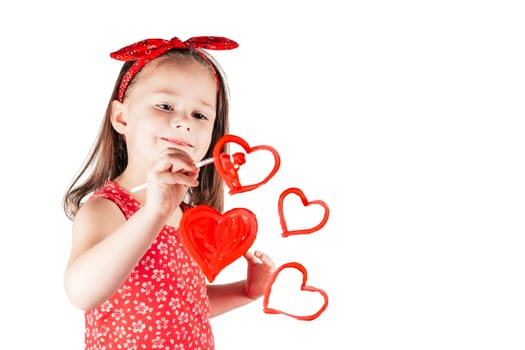 Little girl with long curly hair in red clothes draw hearts with paint on transparent glass smiling isolated on white background. Family, love, holidays, Mother's day concept. Copy space. Mock up.